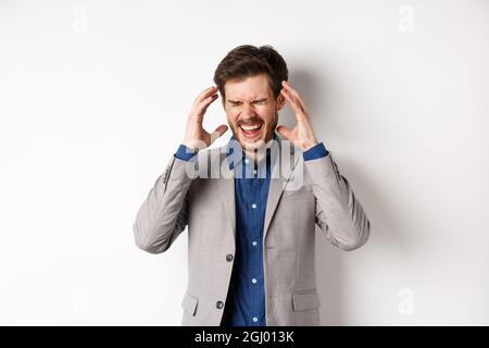 Frustrated business man in suit going crazy, screaming and shaking hands near head, feeling annoyed or angry, shouting distressed, standing on white Stock Photo