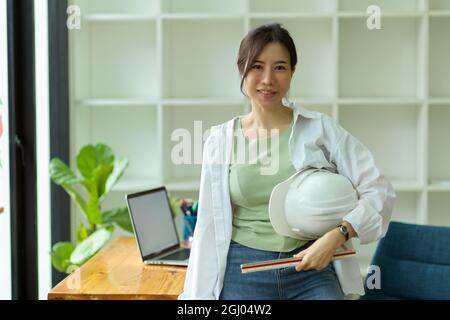 Portrait of young female architect with hardhat and ruler leaning on working desk in modern engineers office Stock Photo