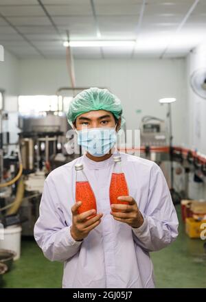 Young asian man worker in sterile uniform showing product of bottled red juice in production line at beverage processing factory Stock Photo