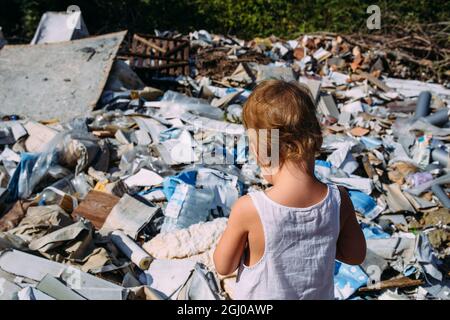 Little girl at a dump among a heap of scattered garbage in the forest. 