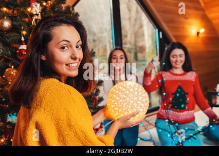 Christmas and new year. Portrait of a young smiling woman holding a lighting ball. In the background, two other women are untangling the lights. Stock Photo