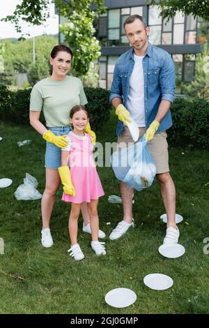Smiling mother hugging daughter in rubber gloves near husband with trash bag outdoors Stock Photo