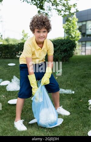 Positive boy in rubber gloves holding bag near trash on grass Stock Photo