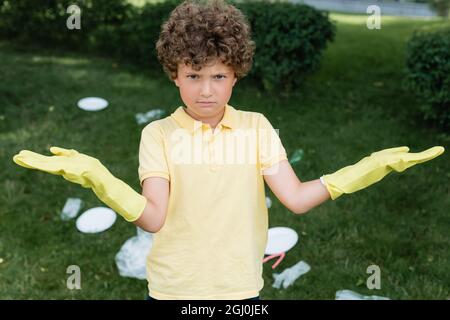 Confused boy in rubber gloves looking at camera near blurred garbage outdoors Stock Photo
