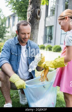 Smiling dad putting bottle in trash bag near daughter on lawn Stock Photo