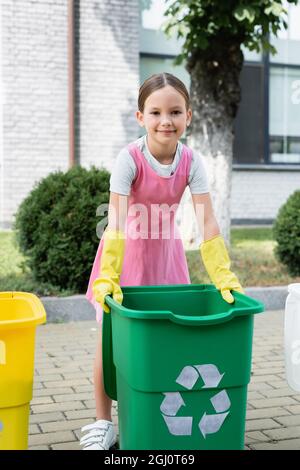 Smiling kid in rubber gloves standing near trash bin with recycle sign outdoors Stock Photo