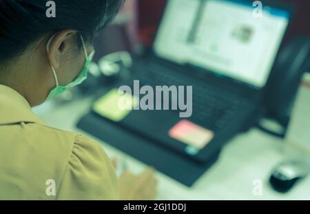 Back view of woman wear face mask working at desk with laptop in office. Asian woman worker using computer at office during work from home. New normal Stock Photo