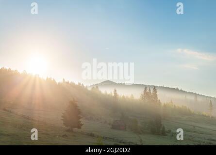 Scenic beautiful panoramic landscape view of old small wooden hut cabin on green mountain hill forest woods meadow at early morning fog misty sunrise Stock Photo
