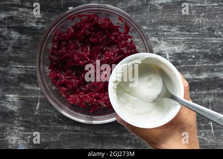 Top view of grated cooked beet in glass bowl and woman hand holding jar with sour cream on black background Stock Photo