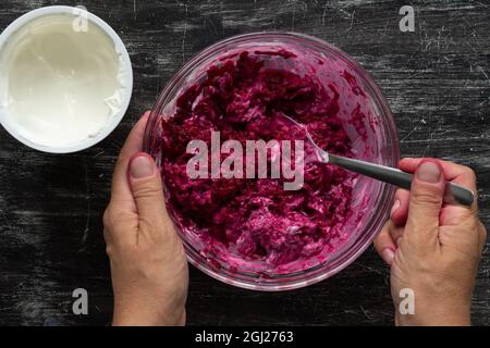 Top view of woman hand mixing grated cooked beet in glass bowl and sour cream with spoon making salad on black background Stock Photo
