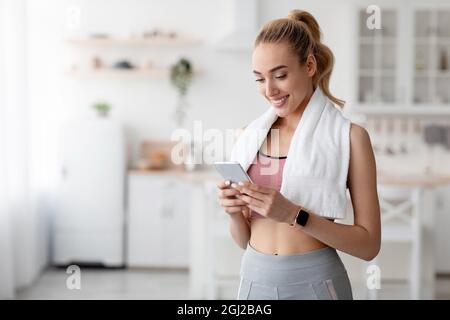 Happy european cute millennial blonde female checking fitness app or social media on smartphone at home, empty space. Sportswoman after exercise, moni Stock Photo