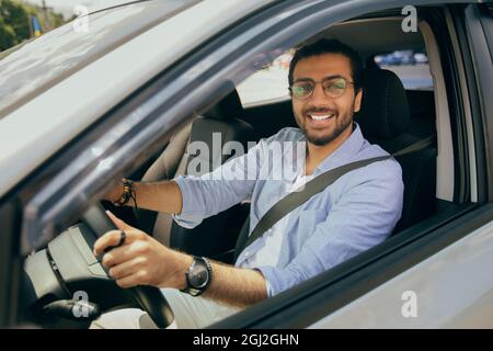 Handsome stylish middle-eastern man driving nice auto, looking through car window and smiling at camera, going to office or travelling alone by automo Stock Photo