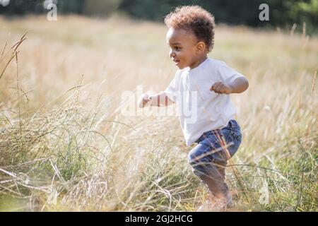 Playful little boy in casual clothes having fun among high grass. Cute african child with curly hair running on field. Freedom and carelessness concept. Stock Photo