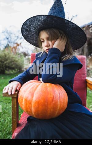 Scary little girl in witch costume, hat with big pumpkin celebrating halloween holiday. Sitting on armchair in coat with pumpkin. Stylish image. Horro Stock Photo