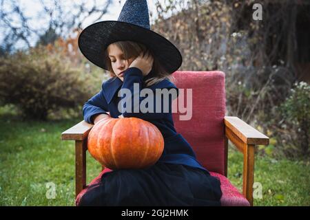 Scary little girl in witch costume, hat with big pumpkin celebrating halloween holiday. Sitting on armchair in coat with pumpkin. Stylish image. Horro Stock Photo