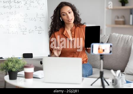 Female math teacher making video conference chat with students using laptop and cellphone on tripod, holding and showing tablet with empty screen with Stock Photo