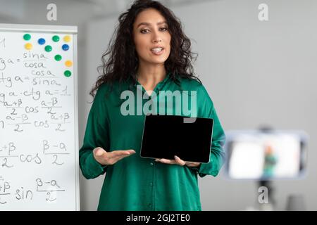 Modern Distance Learning. Female math teacher making video conference chat with students using cellphone on tripod, holding and showing tablet with em Stock Photo