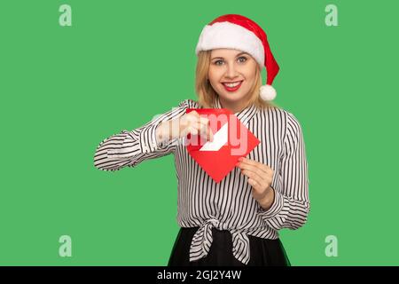 Portrait of blonde young woman in christmas hat standing, holding christmas envelope and looking at camera with toothy smile and enjoying. indoor stud Stock Photo