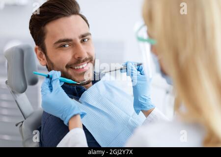Closeup of female dentist blonde woman in workwear doing treatment for patient cheerful young man, holding dental tools, wearing rubber gloves. Stomat Stock Photo