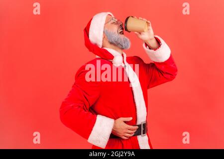 Side view portrait of elderly man with gray beard wearing santa claus costume drinking coffee from paper cup, lack of energy, enjoying hot beverage. I Stock Photo