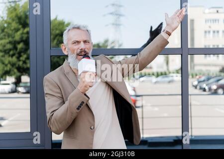 bearded reporter in blazer holding microphone and pointing with hand at building Stock Photo
