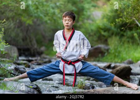 Boy performing martial arts while doing a standing pose on rocks in a forest river. Stock Photo