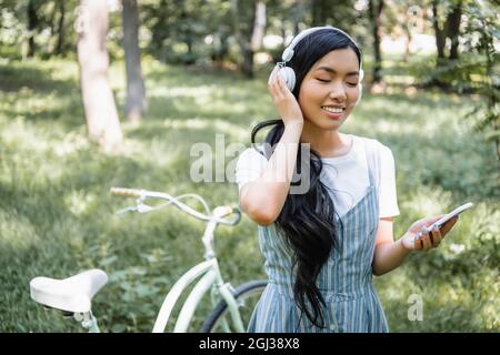 smiling asian woman adjusting headphones while using cellphone near blurred bicycle Stock Photo
