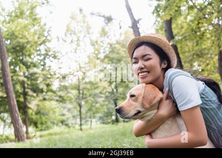 excited asian woman in straw hat embracing labrador in park Stock Photo