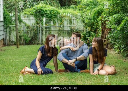 A family of a mother and father and two daughters sitting outside in the grass of their backyard witht the youngest daughter hugging dad from behind Stock Photo