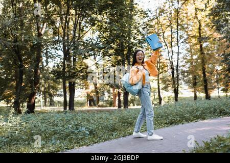 excited asian student waving notebooks while walking in park with backpack Stock Photo