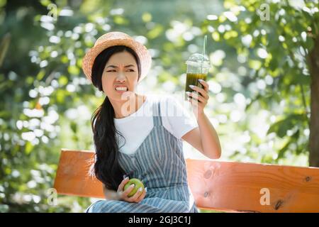 displeased asian woman grimacing while looking at smoothie in plastic cup Stock Photo