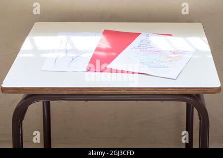 Photograph of some drawings and a red folder of a child on a desk in a classroom of a nursery school.The photo is taken in horizontal format. Stock Photo