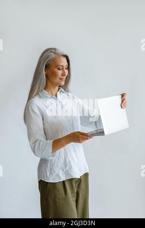 HappyAsian lady with hoary hair opens laptop standing on light grey background Stock Photo