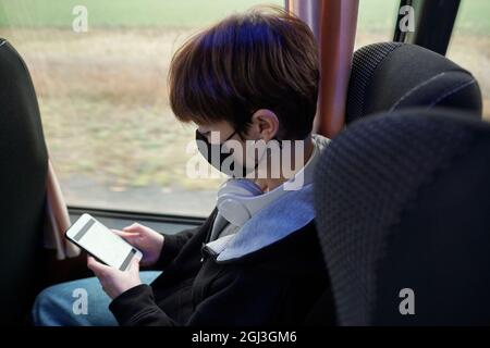 Short-haired young woman in mask wearing white headphones around neck using gadget while riding bus Stock Photo