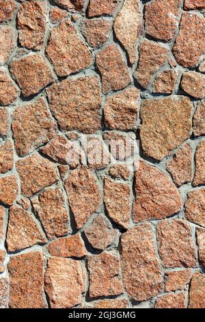 The texture of the red granite wall and the background of large stones embedded in the wall in the light of warm sunlight, close-up, vertical image. Stock Photo
