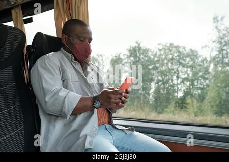 Bald young Black man in mask sitting at window and using smartphone while riding bus Stock Photo