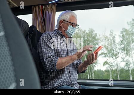 Aged Caucasian man in mask and eyeglasses sitting at window and using phone while sending sms in bus Stock Photo