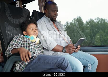 Afro American boy with headphones around neck sleeping on fathers shoulder in bus while he using tablet Stock Photo