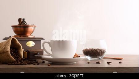 Cup of hot coffee with sack full of beans and manual grinder on wooden table and white isolated background. Front view. Horizontal composition. Stock Photo