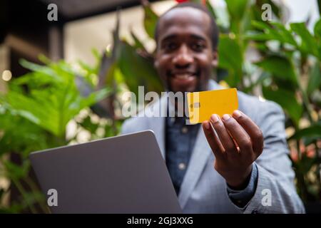 African businessman is holding a credit card and is making an online purchase with his laptop. Smiling elegant millennial man - technological man buyi Stock Photo