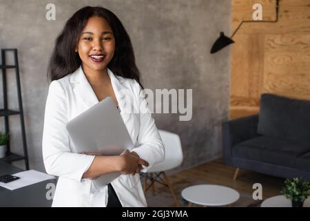 Handsome african american business woman in classic suit uses laptop and smiles while working in office Stock Photo