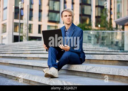 An office worker with laptop on his knees sits on step in front of building of business center. Stock Photo