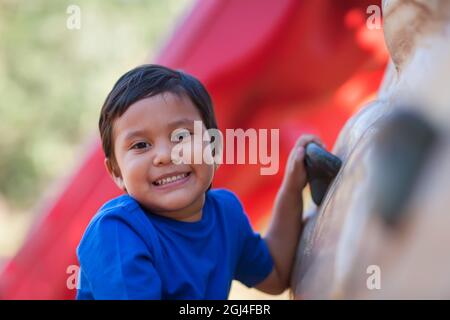 5 year old boy smiles while holding on to a kids outdoor rock climbing wall. Stock Photo