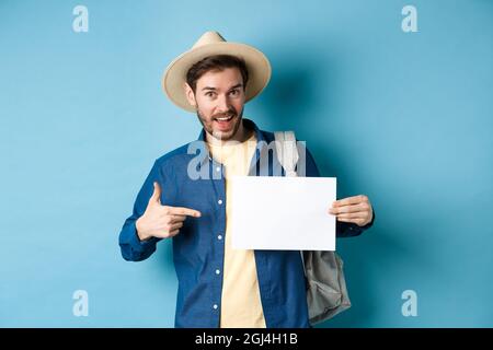 Handsome tourist smiling and pointing at empty piece of paper, wearing straw hat and backpack. Concept of summer vacation Stock Photo