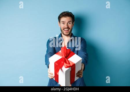 Valentines day. Excited handsome man giving gift box and wishing happy holiday. Romantic guy extending hands with present, standing againt blue Stock Photo