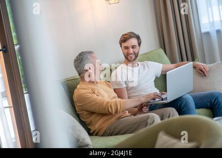 Young bearded man working on a laptop, his father sitting next to him Stock Photo