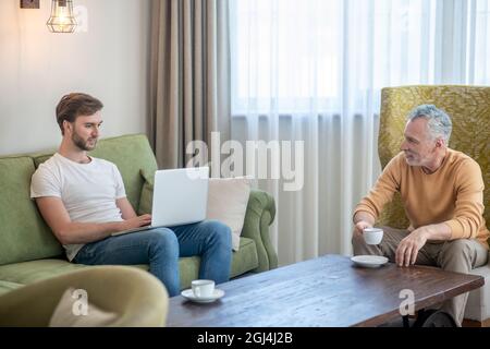 Young bearded man working on a laptop, his father drinking tea Stock Photo