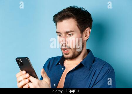 Close-up of man looking shocked and reading message on smartphone, gasping and stare startled at screen, standing on blue background Stock Photo