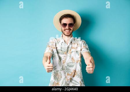 Happy tourist in sunglasses and straw hat showing thumbs up and smiling, enjoying summer holiday, standing on white background Stock Photo