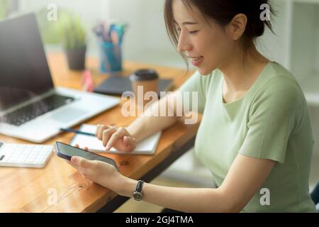 Side view, Casual businesswoman using smartphone on working desk in office, On desk are laptop, notebook, stationery, take away coffee Stock Photo
